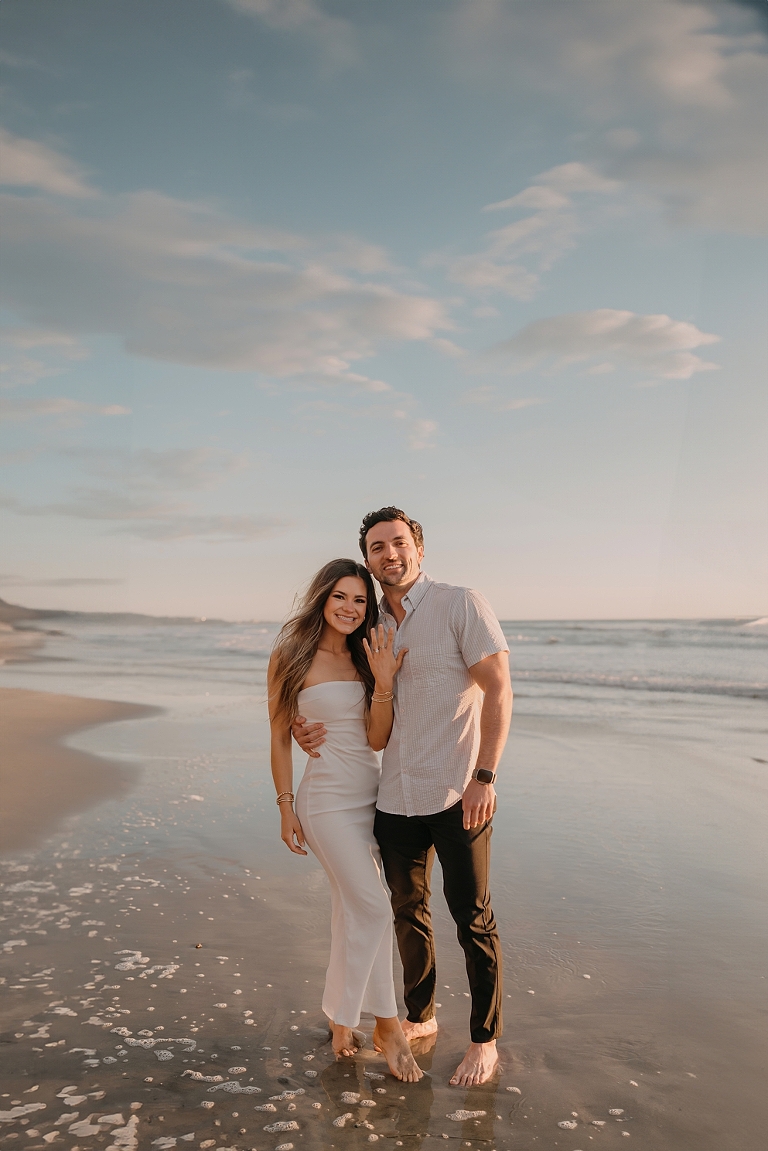 San Diego Proposal Photographer A couple standing closely on Del Mar Beach at sunset, smiling towards the camera, with waves softly breaking near their feet.